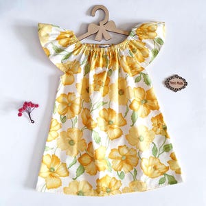May include: A white dress with a yellow floral pattern and ruffled sleeves hangs on a wooden hanger. The dress features a scoop neckline and a flared skirt. A small wooden sign that says "Hand Made" is next to the dress.