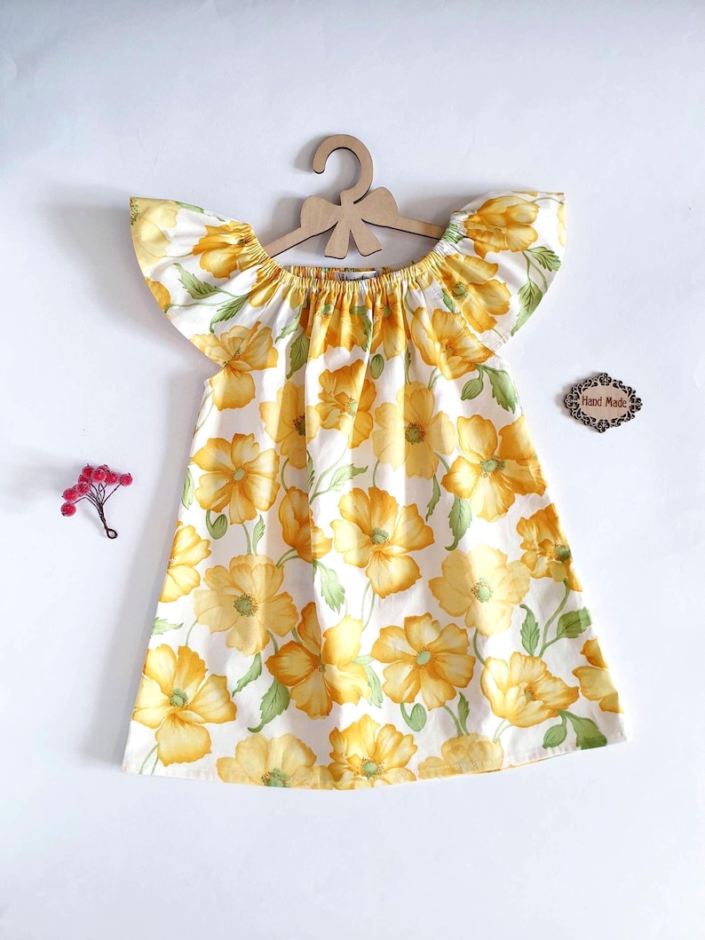 May include: A white dress with a yellow floral pattern and ruffled sleeves hangs on a wooden hanger. The dress features a scoop neckline and a flared skirt. A small wooden sign that says "Hand Made" is next to the dress.