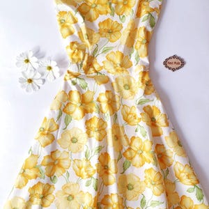 May include: A sleeveless, white dress with a vibrant yellow floral pattern. The dress features a V-neckline and a fitted bodice that flows into a flared skirt. The dress is adorned with large yellow flowers and green leaves. The words "Hand Made" are on a small wooden tag.