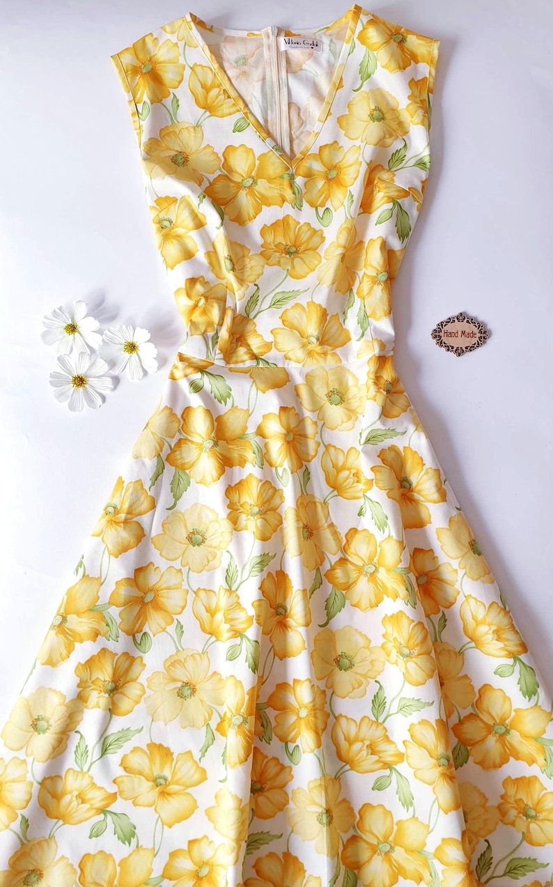 May include: A sleeveless, white dress with a vibrant yellow floral pattern. The dress features a V-neckline and a fitted bodice that flows into a flared skirt. The dress is adorned with large yellow flowers and green leaves. The words "Hand Made" are on a small wooden tag.