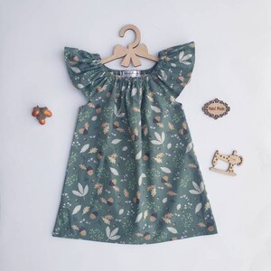 May include: A green dress with a leaf and acorn pattern, displayed on a wooden hanger. The dress has flutter sleeves and a gathered neckline. Decorative wooden cutouts of a sewing machine and a sign that says "Hand Made" are also visible.