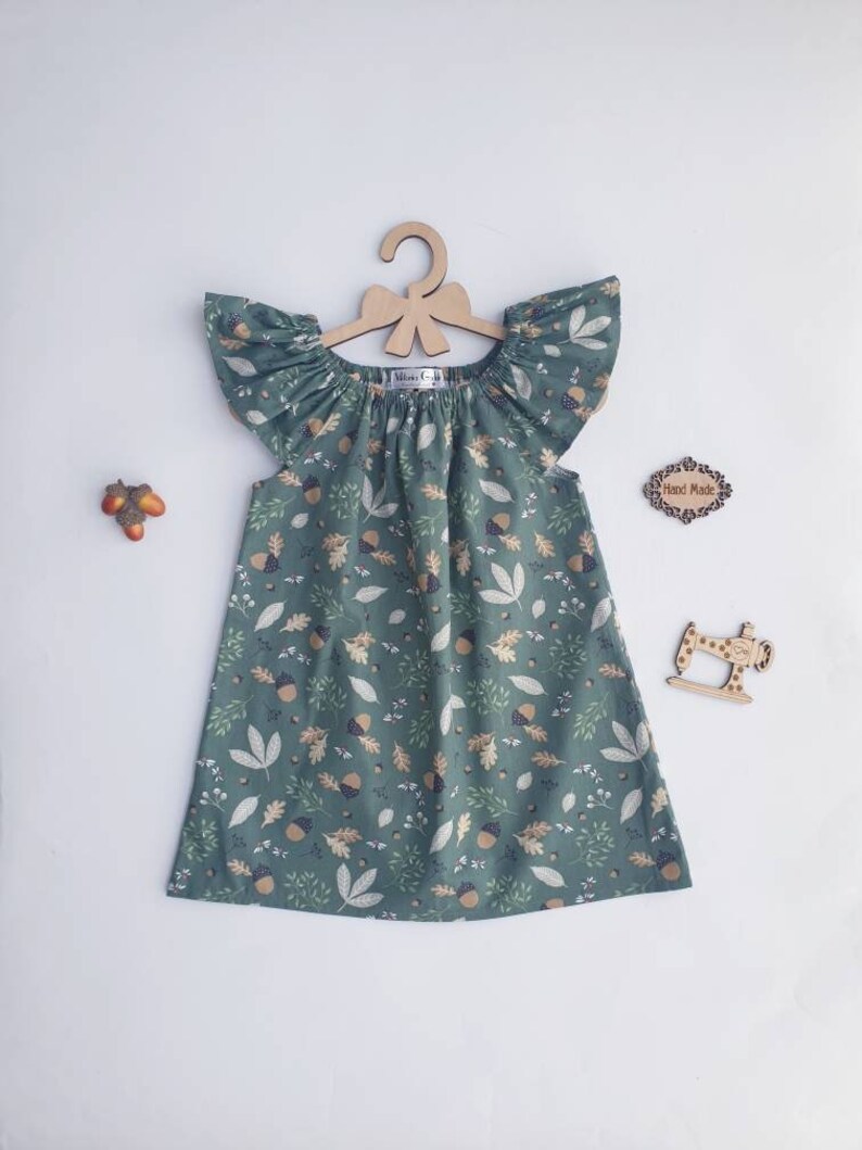 May include: A green dress with a leaf and acorn pattern, displayed on a wooden hanger. The dress has flutter sleeves and a gathered neckline. Decorative wooden cutouts of a sewing machine and a sign that says "Hand Made" are also visible.