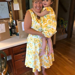 May include: A woman and a child are embracing, both wearing matching yellow dresses with a floral pattern. The dresses feature large yellow flowers on a white background. The scene is indoors, with a wooden floor and cabinets visible.