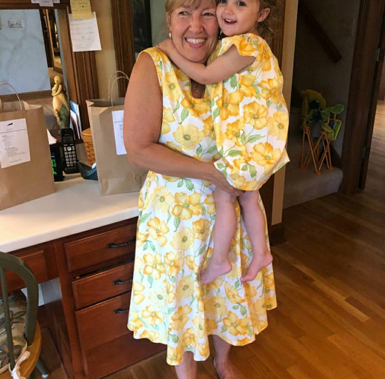 May include: A woman and a child are embracing, both wearing matching yellow dresses with a floral pattern. The dresses feature large yellow flowers on a white background. The scene is indoors, with a wooden floor and cabinets visible.