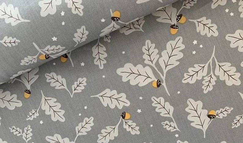May include: Gray fabric with a repeating pattern of white oak leaves, acorns, and small white stars. The acorns have yellow caps and brown details. The leaves have black veins. The fabric is folded over, showing the pattern in a layered effect.