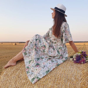 K&ouml;nnte beinhalten: Eine Frau in einem wei&szlig;en Blumenkleid und einem wei&szlig;en Hut sitzt auf einem Heuballen auf einem Feld. Sie h&auml;lt einen Strau&szlig; lila und wei&szlig;er Blumen.