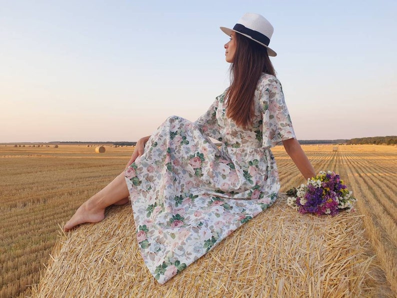 K&ouml;nnte beinhalten: Eine Frau in einem wei&szlig;en Blumenkleid und einem wei&szlig;en Hut sitzt auf einem Heuballen auf einem Feld. Sie h&auml;lt einen Strau&szlig; lila und wei&szlig;er Blumen.