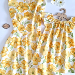 May include: A matching set of dresses with a floral pattern. The dresses are white with large yellow flowers and green leaves. One dress has a V-neck and the other has a ruffled neckline. The dresses are displayed on a white background.