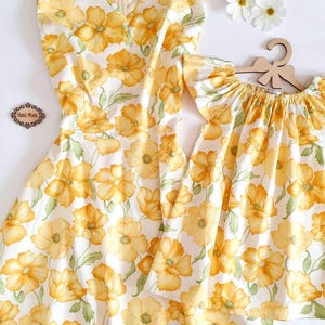 May include: A sleeveless dress and a matching top for a child, both featuring a white background with a repeating pattern of large yellow flowers and green leaves. The dress has a V-neck and a zipper. The child's top has ruffled sleeves.
