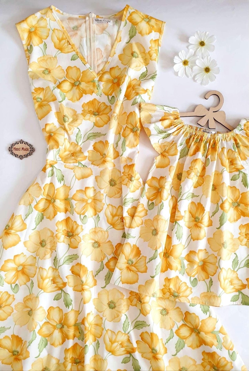 May include: A sleeveless dress and a matching top for a child, both featuring a white background with a repeating pattern of large yellow flowers and green leaves. The dress has a V-neck and a zipper. The child's top has ruffled sleeves.