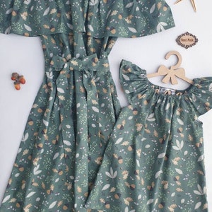 May include: Matching green dresses with an off-the-shoulder design and a leaf and acorn pattern. One dress is full-length with a waist tie, and the other is a shorter, flutter-sleeve style. A wooden hanger and starfish are also visible.