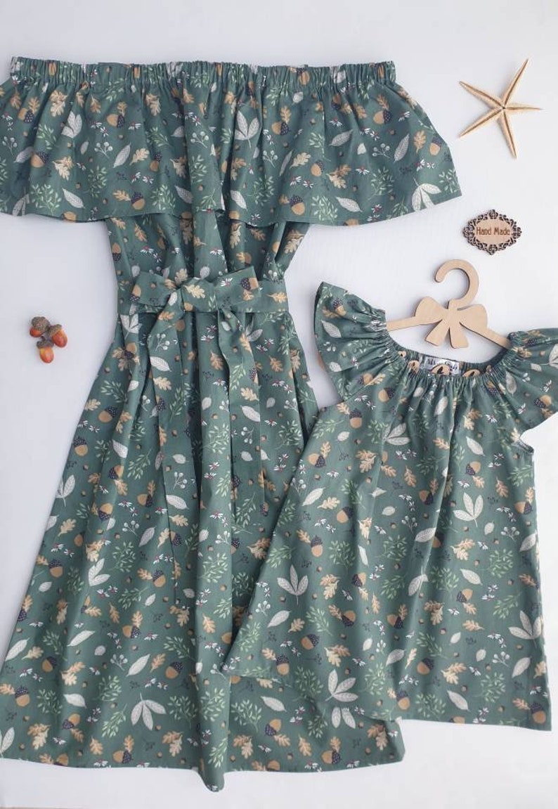 May include: Matching green dresses with an off-the-shoulder design and a leaf and acorn pattern. One dress is full-length with a waist tie, and the other is a shorter, flutter-sleeve style. A wooden hanger and starfish are also visible.