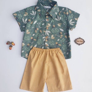 May include: A two-piece children's outfit featuring a short-sleeved button-down shirt and matching shorts. The shirt is a muted green with a pattern of leaves and acorns. The shorts are mustard yellow with a repeating chevron design. The outfit is handmade.