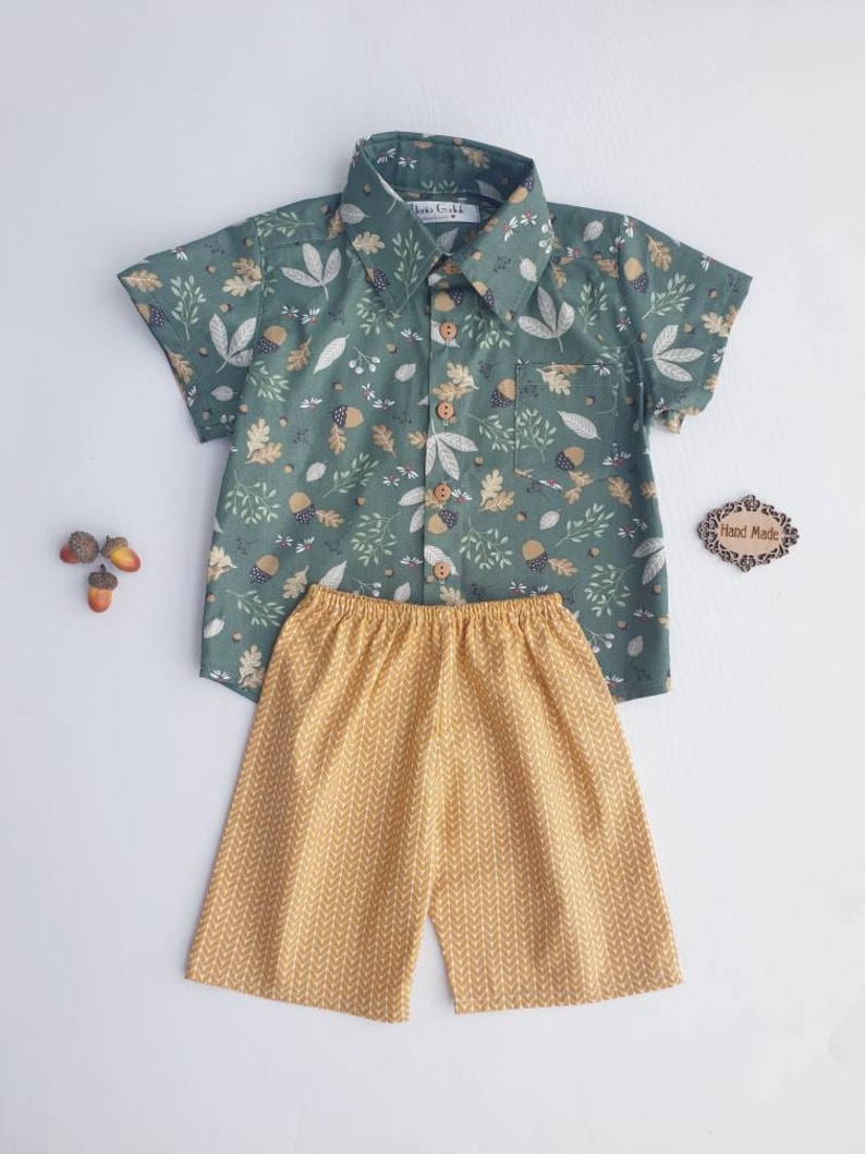 May include: A two-piece children's outfit featuring a short-sleeved button-down shirt and matching shorts. The shirt is a muted green with a pattern of leaves and acorns. The shorts are mustard yellow with a repeating chevron design. The outfit is handmade.