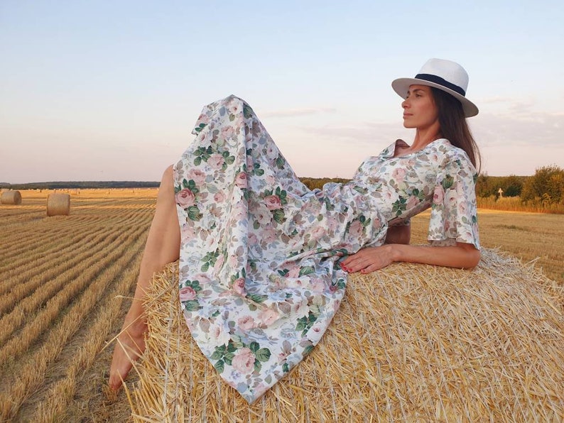 K&ouml;nnte beinhalten: Eine Frau in einem wei&szlig;en Hut und einem Blumenkleid liegt auf einem Heuballen auf einem Feld. Das Kleid hat einen wei&szlig;en Hintergrund mit rosa und gr&uuml;nen Blumenmustern.