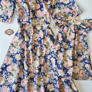 K&ouml;nnte beinhalten: Ein passendes Set aus zwei Blumenkleidern auf dunkelblauem Grund mit rosa, wei&szlig;en und gelben Blumen. Das gr&ouml;&szlig;ere Kleid hat einen V-Ausschnitt und kurze &Auml;rmel. Das kleinere Kleid hat einen quadratischen Ausschnitt und kurze, gekr&auml;uselte &Auml;rmel.