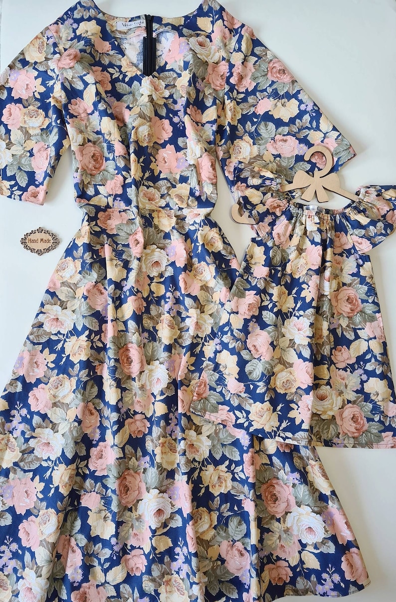 K&ouml;nnte beinhalten: Ein passendes Set aus zwei Blumenkleidern auf dunkelblauem Grund mit rosa, wei&szlig;en und gelben Blumen. Das gr&ouml;&szlig;ere Kleid hat einen V-Ausschnitt und kurze &Auml;rmel. Das kleinere Kleid hat einen quadratischen Ausschnitt und kurze, gekr&auml;uselte &Auml;rmel.