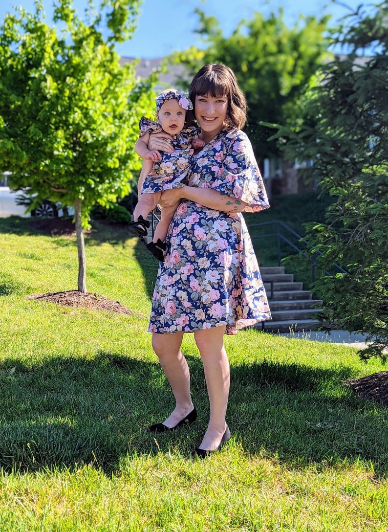 K&ouml;nnte beinhalten: Eine Frau und ein kleines M&auml;dchen tragen passende Blumenkleider. Die Kleider sind blau mit rosa und wei&szlig;en Blumen. Die Frau h&auml;lt das M&auml;dchen in ihren Armen. Beide l&auml;cheln und schauen in die Kamera.