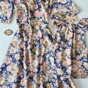 K&ouml;nnte beinhalten: Zwei Blumenkleider, eines lang und eines kurz, mit blauem Hintergrund und rosa, gelben und wei&szlig;en Blumen. Die Kleider haben einen V-Ausschnitt und kurze &Auml;rmel. Der Text "Hand Made" ist auf einem kleinen Etikett sichtbar.
