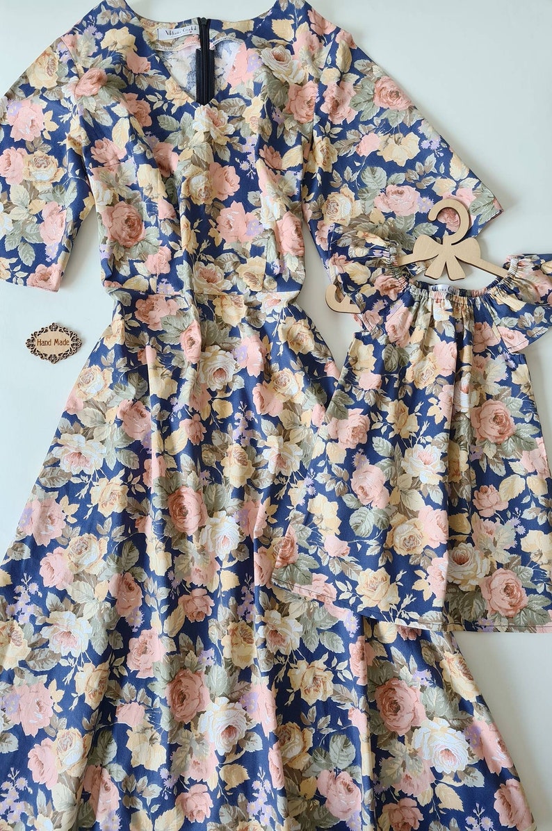 K&ouml;nnte beinhalten: Zwei Blumenkleider, eines lang und eines kurz, mit blauem Hintergrund und rosa, gelben und wei&szlig;en Blumen. Die Kleider haben einen V-Ausschnitt und kurze &Auml;rmel. Der Text "Hand Made" ist auf einem kleinen Etikett sichtbar.