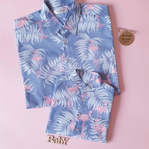 Peut inclure: Deux chemises boutonnées bleues avec un imprimé de flamants roses et de feuilles de palmier blanches. La plus grande chemise a un col classique, tandis que la plus petite a un col officier. Une étiquette en bois indique "HAND MADE with love" et un panneau en bois indique "Baby".