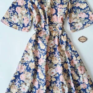 K&ouml;nnte beinhalten: Ein blaues Blumenkleid mit V-Ausschnitt und kurzen &Auml;rmeln. Das Kleid hat ein tailliertes Oberteil und einen flie&szlig;enden Rock. Der Stoff ist eine leichte Baumwolle mit einem Blumendruck in Rosa-, Gelb- und Gr&uuml;nt&ouml;nen. Das Kleid hat einen Rei&szlig;verschluss auf der R&uuml;ckseite. Das Kleid ist handgefertigt.