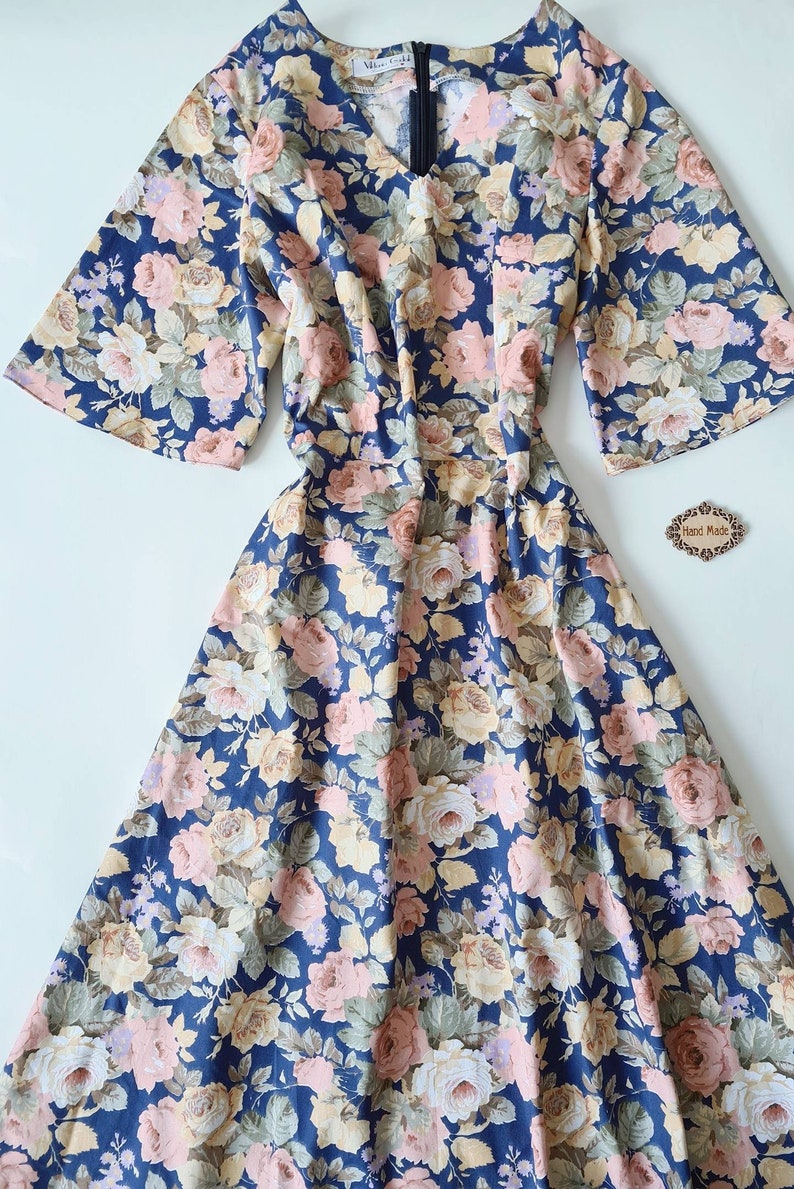 K&ouml;nnte beinhalten: Ein blaues Blumenkleid mit V-Ausschnitt und kurzen &Auml;rmeln. Das Kleid hat ein tailliertes Oberteil und einen flie&szlig;enden Rock. Der Stoff ist eine leichte Baumwolle mit einem Blumendruck in Rosa-, Gelb- und Gr&uuml;nt&ouml;nen. Das Kleid hat einen Rei&szlig;verschluss auf der R&uuml;ckseite. Das Kleid ist handgefertigt.