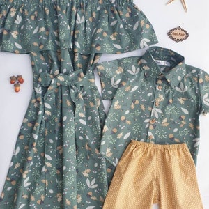 May include: A green off-the-shoulder dress with a matching short-sleeved button-up shirt and yellow shorts. All items feature a leaf and acorn pattern. A starfish and a "Hand Made" tag are also present.