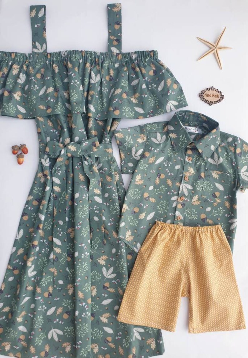 May include: A green off-the-shoulder dress with a matching short-sleeved button-up shirt and yellow shorts. All items feature a leaf and acorn pattern. A starfish and a "Hand Made" tag are also present.