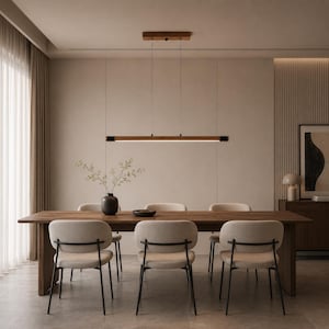 May include: A linear wooden pendant light fixture suspended above a long rectangular wooden dining table. Six upholstered chairs with black metal frames are arranged around the table. A dark ceramic vase with branches sits on the table surface.