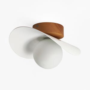 May include: A ceiling light fixture featuring a circular wooden base with a visible screw, a wide white curved metal shade, and a central spherical frosted glass globe. The design combines natural wood grain with smooth white metal and glass.