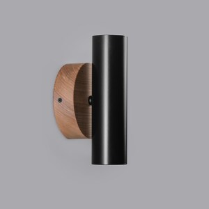Puede incluir: Aplique de pared moderno con una l&aacute;mpara cil&iacute;ndrica negra fijada a una base de madera circular. La madera tiene un patr&oacute;n de vetas natural. El aplique est&aacute; dise&ntilde;ado para iluminaci&oacute;n interior.