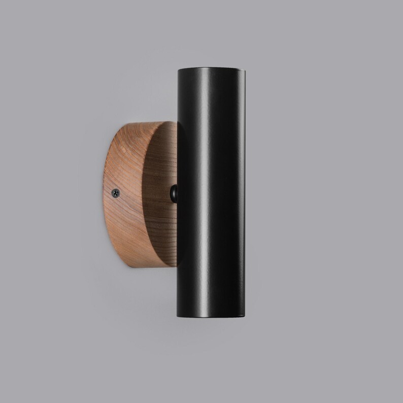 Puede incluir: Aplique de pared moderno con una l&aacute;mpara cil&iacute;ndrica negra fijada a una base de madera circular. La madera tiene un patr&oacute;n de vetas natural. El aplique est&aacute; dise&ntilde;ado para iluminaci&oacute;n interior.