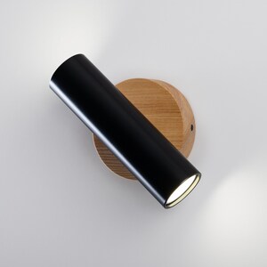 Puede incluir: Un foco de pared moderno. El accesorio presenta un cabezal de luz cil&iacute;ndrico negro fijado a una base redonda de madera de color claro. La luz est&aacute; en &aacute;ngulo y emite un haz brillante y enfocado. Dise&ntilde;o minimalista y contempor&aacute;neo.