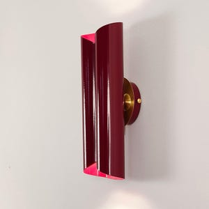 May include: A vertical wall sconce featuring a glossy deep burgundy metal shade with a curved, dual-panel design. The interior of the shade reveals a bright pink finish. A circular brass-toned mounting plate connects the fixture to the wall.