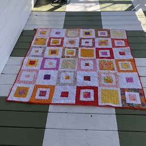 May include: A colorful patchwork quilt with squares of orange, yellow, pink, and purple fabric. The quilt is laid out on a wooden porch floor.