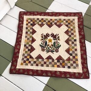 May include: A small, cream-colored quilt with a red border and a brown, green, and gold applique design in the center. The design features a wreath of leaves, acorns, and berries with a gold star in the center.