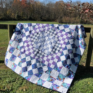 May include: A handmade quilt with a geometric pattern of white, purple, and blue squares. The quilt is draped over a wooden fence in an outdoor setting, showcasing its intricate design and craftsmanship. The quilt's colors and pattern create a visually appealing textile.