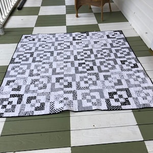 May include: Black and white quilt with a geometric pattern. The quilt is made up of squares of different sizes and patterns, all in black and white. The quilt is lying on a checkered floor.