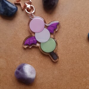 Peut inclure: Un charme dango violet et vert avec un motif d'aile de chauve-souris violette. Le charme est attaché à un porte-clés en forme d'étoile dorée.