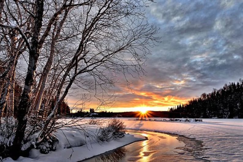 May include: A snowy winter landscape with a frozen river winding through it. The sun is setting behind a line of trees, casting a warm glow over the scene.