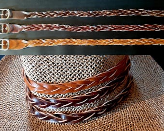May include: Three braided leather belts in varying shades of brown, ranging from light tan to dark chocolate. Each belt features a silver-toned metal buckle and a smooth leather end piece. The belts are displayed both laid flat and coiled.