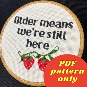 May include: A white embroidery hoop with a cross-stitch design that reads "Older means we're still here" with two red strawberries on a green vine.