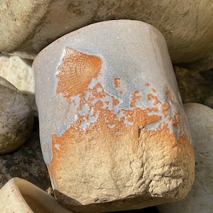 May include: A ceramic mug with a light grey glaze and a brown, textured base. The glaze has a mottled appearance, with areas of light and dark grey.