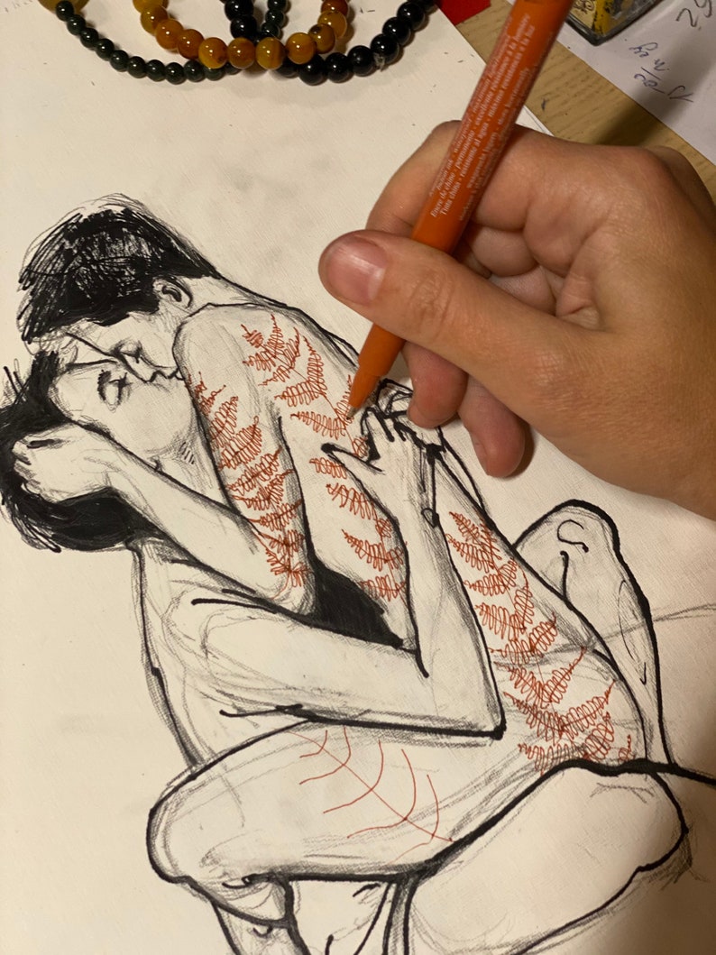 May include: A black ink line drawing on white paper depicting two figures in an embrace. One figure features intricate red fern-like patterns drawn along the back and leg. An orange marker is shown in the process of adding these red details.