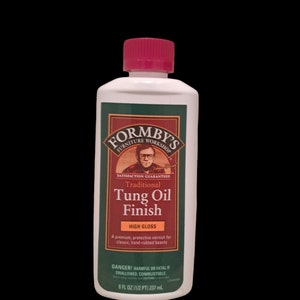May include: A bottle of Formby's Furniture Workshop Traditional Tung Oil Finish in High Gloss. The bottle is green and white with a red cap. It features a picture of a man with glasses and a mustache. The bottle is 8 fluid ounces (237 ml).