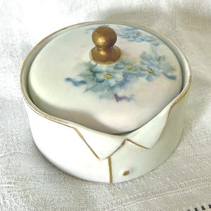 May include: A white porcelain trinket box with a lid featuring a floral design in shades of blue and green. The lid is topped with a gold-colored knob. The box has gold trim and a unique collar-like shape.