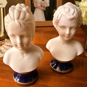 May include: Two white matte porcelain busts of children, each featuring detailed facial features and hair. The busts are mounted on circular, deep blue glazed ceramic bases with gold-toned metallic trim along the bottom edges.