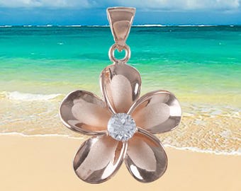 Puede incluir: Un colgante de flor de plumeria de oro rosa con un centro de cristal transparente. El colgante está en una playa de arena blanca con agua de mar azul en el fondo.