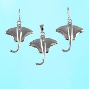 Op de afbeelding: Drie zilveren roggen hangers, elk met een gedetailleerd ontwerp van een rog met zijn vleugels uitgespreid. De hangers hangen tegen een lichtblauwe achtergrond.